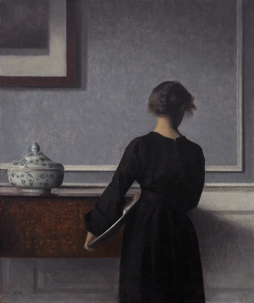 Vilhelm Hammershoi, Interior with Young Woman Seen from the Back (1903-04): a young woman in a dark dress stands with her back to the viewer in a quiet grey interior.